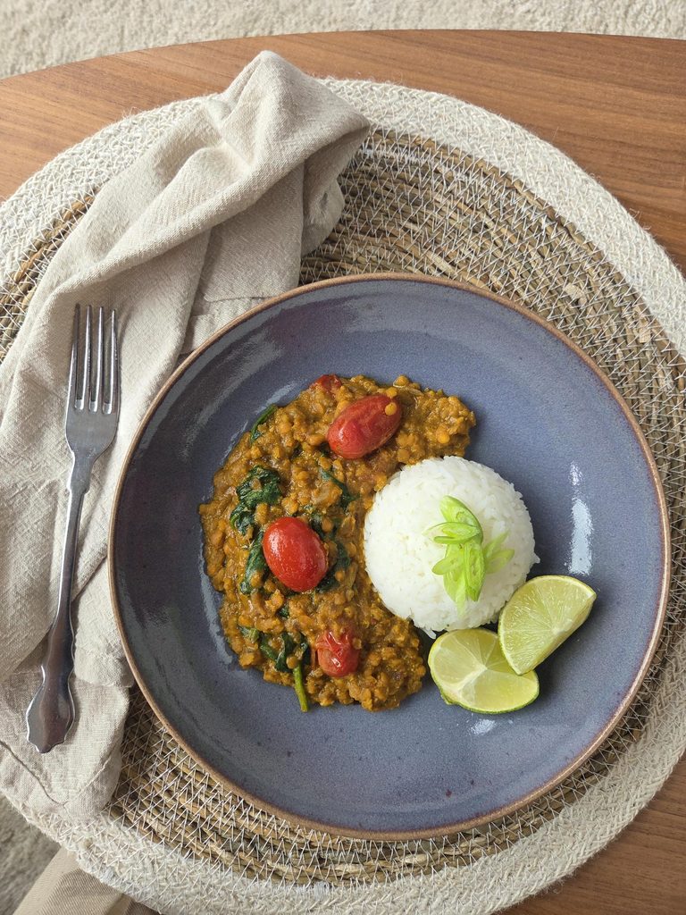 Dal de lentilles corail aux épinards et tomates cerises, servi avec un dôme de riz, de la cébette et du citron vert, dans une assiette bleue sur un set en fibres naturelles.