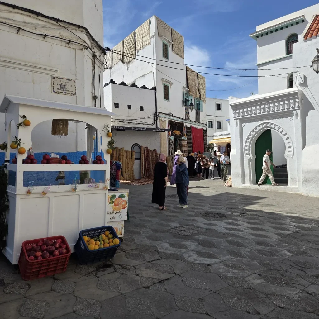 Ruelle blanchie à la chaux de la médina d’Asilah, au Maroc, avec un étal de jus d’orange et de grenades et une porte voûtée verte.